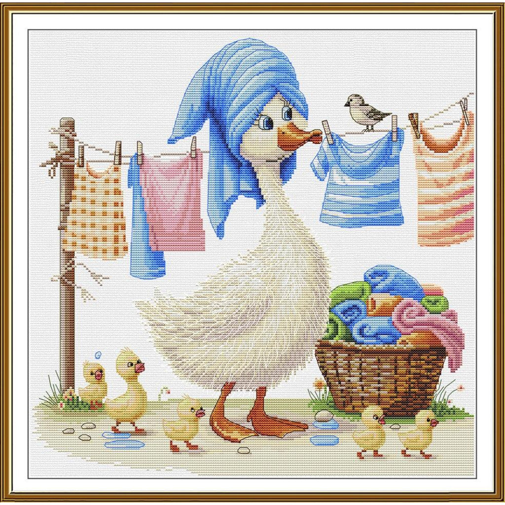 A framed needlepoint or cross-stitch style artwork showing a white goose with a blue towel wrapped around her neck, orange feet, and several yellow goslings around her. Laundry hangs on a line, and a basket of folded towels sits on the ground.