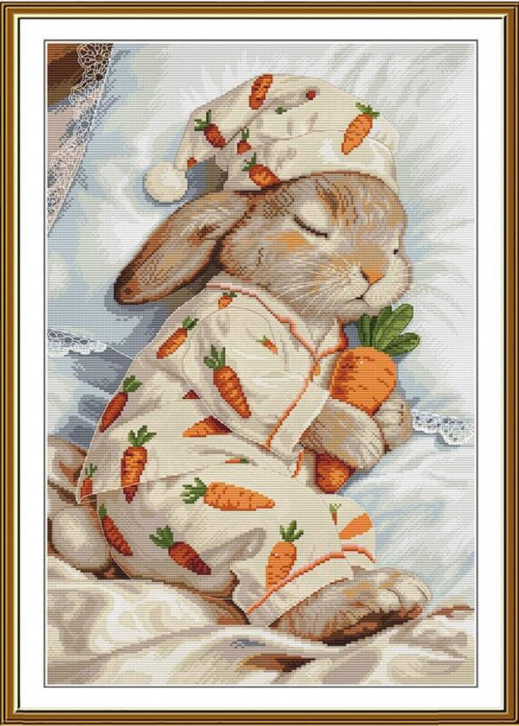 A framed needlepoint or cross-stitch style artwork showing a sleeping rabbit wearing a cream pajama set with an orange carrot print and a matching hat, holding an orange carrot, with a gold-toned frame and white mat border.