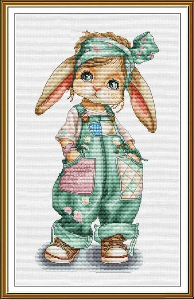 A framed cross-stitch style illustration of a cute bunny character wearing green overall-style clothing, a matching head bow, and brown shoes, shown against a white background with a gold-toned frame.