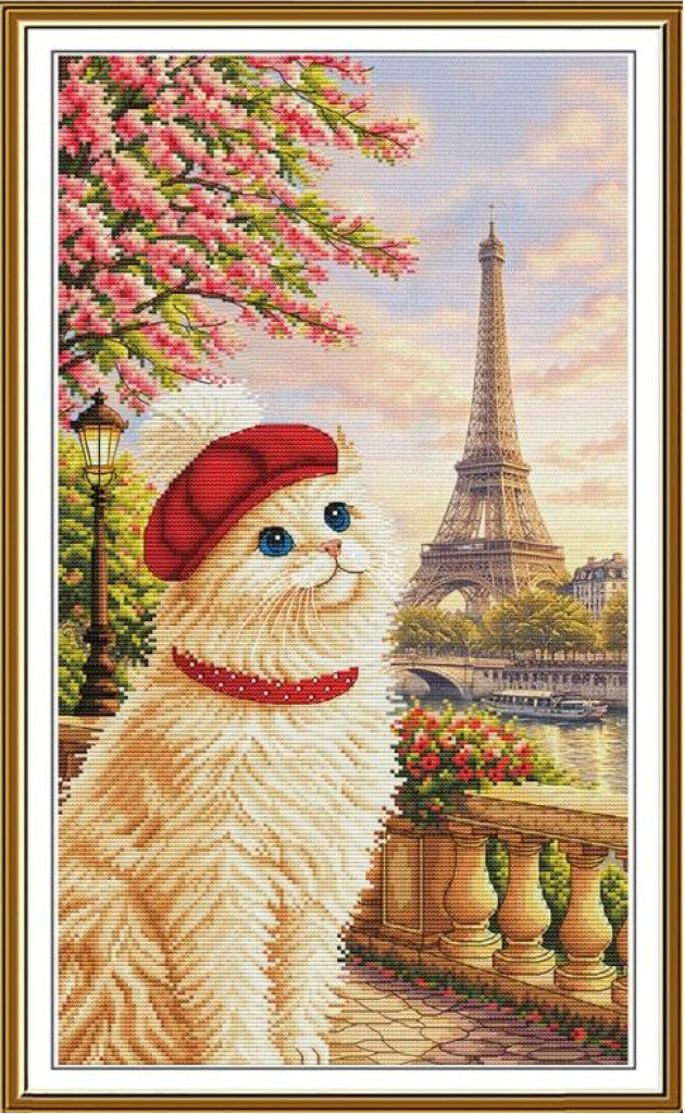 A framed cross-stitch style artwork showing a fluffy cream-colored cat wearing a red beret and red collar, sitting beside a Parisian balcony with the Eiffel Tower and a river in the background. Pink blossoms hang from a tree on the left, and the scene is enclosed in a decorative gold-toned frame.