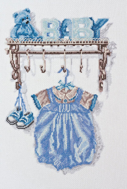 Cross stitch kit depicting a baby-themed scene with a blue dress, shoes, and the word 'BABY' in block letters, all designed in a stitched format on unprinted canvas.