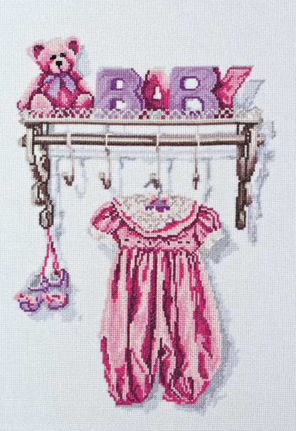 Cross stitch kit depicting a baby girl's outfit with the word 'BABY' and a teddy bear hanging on display.