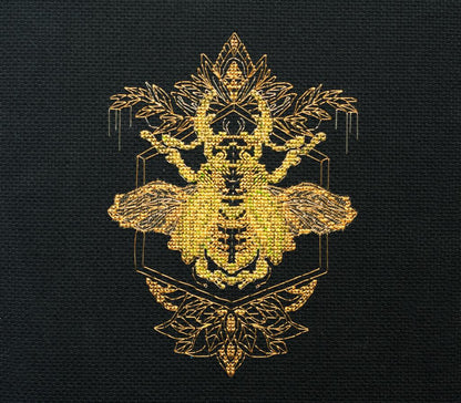 A cross stitch kit depicting a golden beetle on a black background, with intricate gold patterns around the insect.