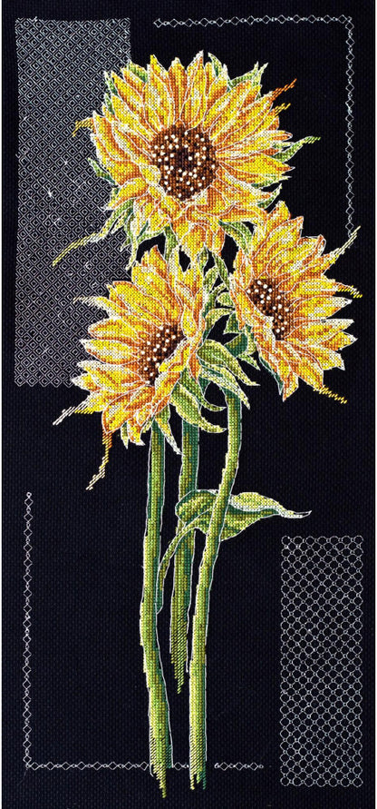 A cross stitch kit depicting three sunflowers with black background, kit contents visible including threads, fabric, a symbol chart, needle, and instructions.