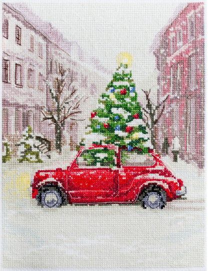 Counted cross stitch kit depicting a red car delivering a Christmas tree, set against a snowy street and building background.