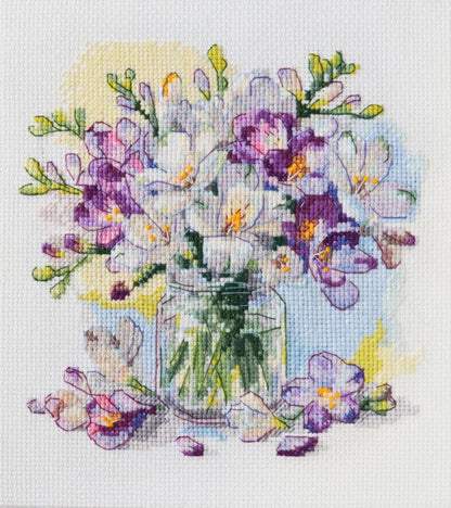 A completed counted cross stitch kit depicting a bouquet of purple and white flowers in a clear vase, set against a light background.