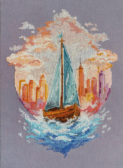 A completed counted cross stitch kit depicting a sailboat on a body of water with a city skyline in the background, stitched on Aida fabric with a needle and thread.