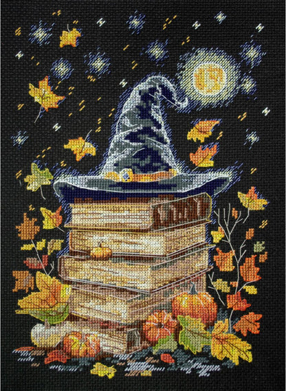 Cross stitch kit image depicting a stack of books with a wizard's hat on top, surrounded by autumn leaves and a moon in the background.