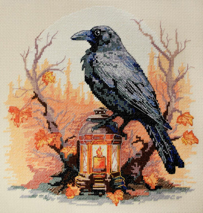 A cross stitch kit depicting a black raven perched on a lantern with autumn leaves and trees in the background.