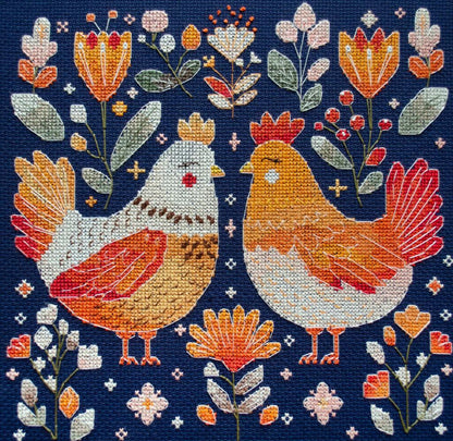 A completed counted cross stitch kit depicting two chickens among floral designs on a dark background.