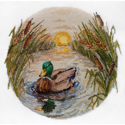 A completed cross stitch kit depicting a duck in water surrounded by reeds with a sunset in the background.