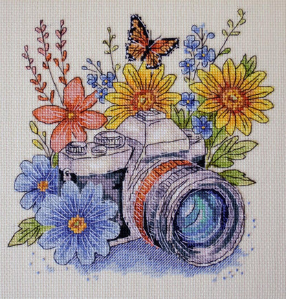 Cross-stitched design of a camera surrounded by flowers and a butterfly on a white background.