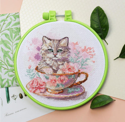 Cross-stitched picture of a cat in a teacup with green leaves in the background