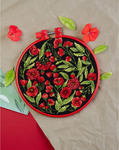 A completed counted cross stitch kit depicting red flowers surrounded by green leaves, displayed in a red hoop.