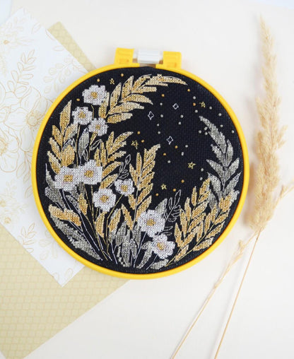 A cross stitch kit depicting white and yellow flowers on a black fabric background, displayed in a yellow hoop, with beige fabric and a wheat stem in the background.