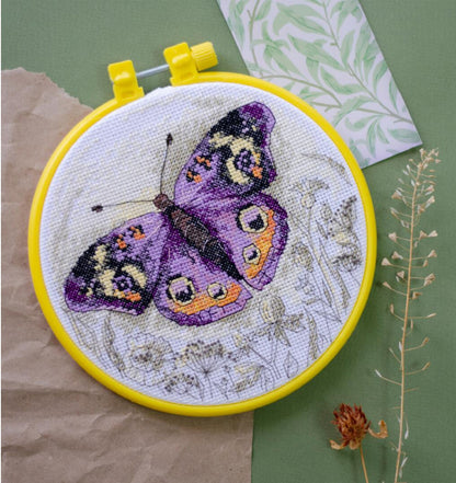A cross stitch kit depicting a purple butterfly on a yellow background, with green fabric and a needle included.