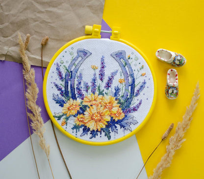 Counted cross stitch kit by Abris Art depicting a floral horseshoe design, displayed on a yellow surface with a decorative hoop and various crafting materials around it.
