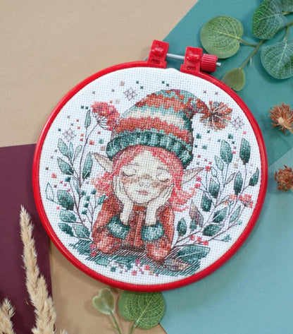 A cross stitch kit depicting a dreaming elf, surrounded by decorative elements like leaves and snowflakes, displayed in a red hoop.
