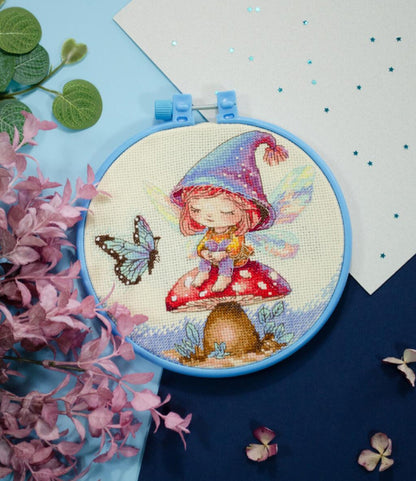 A cross stitch kit depicting a floral gnome design with a decorative hoop, fabric, and threads displayed on a table.