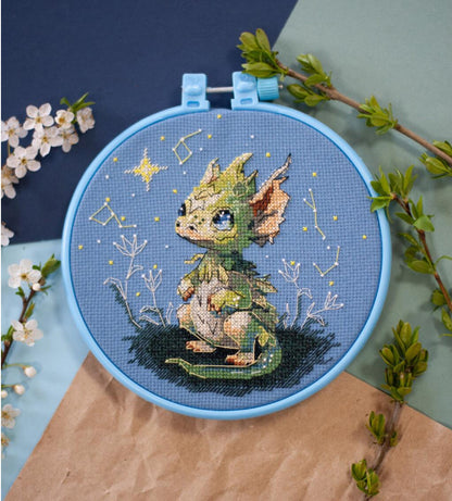 A cross stitch kit depicting a fantasy little dragon with a decorative hoop, surrounded by floral elements and a starry background.