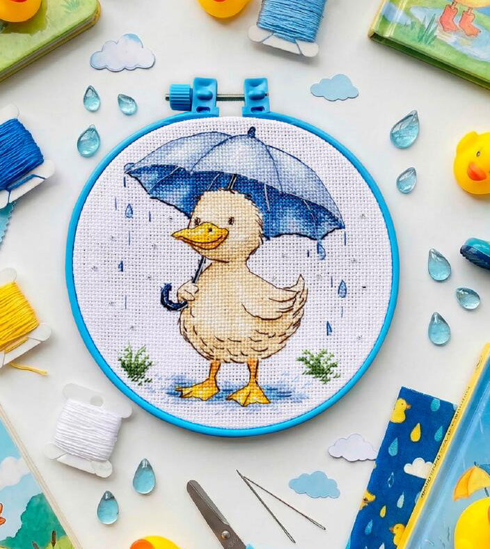 Cross-stitched duck holding an umbrella in a hoop, surrounded by sewing supplies on a light background.