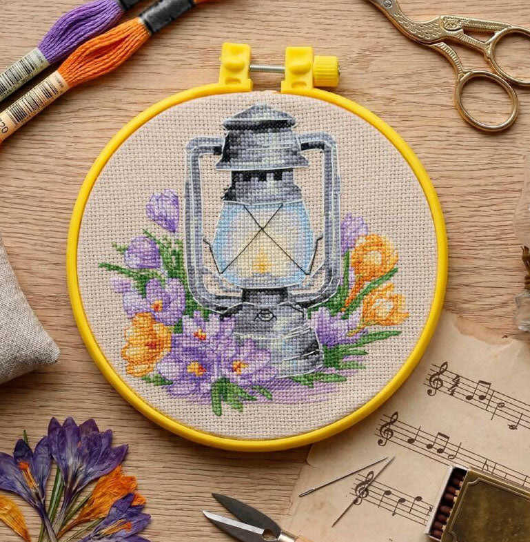 Embroidery hoop with a lantern and floral design on a wooden surface with embroidery tools.