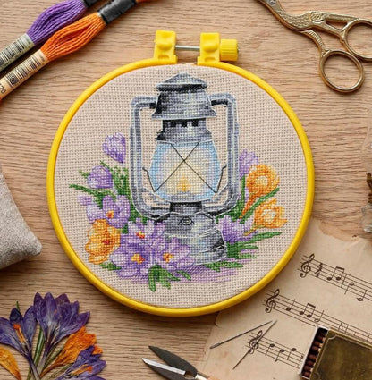 Embroidery hoop with a lantern and floral design on a wooden surface with embroidery tools.