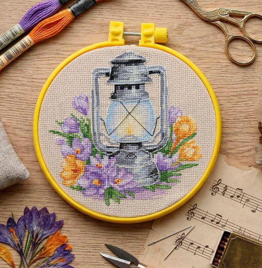 Embroidery hoop with a lantern and floral design on a wooden surface with embroidery tools.