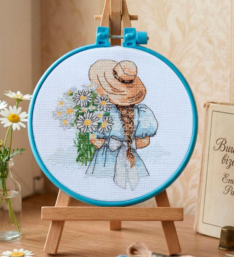 Embroidery hoop with a cross-stitched design of a girl with flowers on a wooden stand.