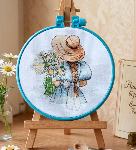 Embroidery hoop with a cross-stitched design of a girl with flowers on a wooden stand.