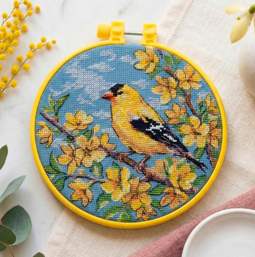 Embroidery hoop with a yellow bird and floral design on a blue background, surrounded by yellow flowers and green leaves.