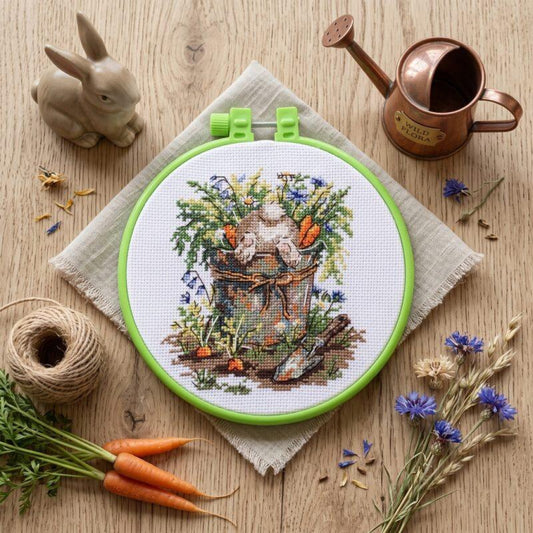 A cross-stitch embroidery hoop displaying a bunny in a garden basket, styled on a wooden surface with a small rabbit figurine, copper watering can, twine, carrots, and wildflowers.