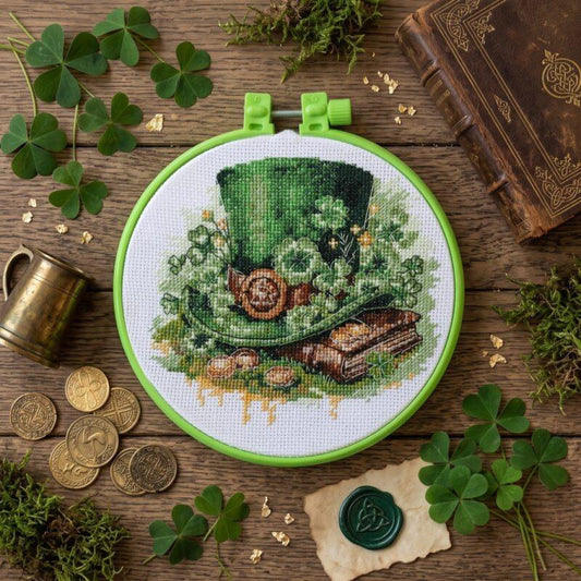 A circular embroidery hoop containing a stitched design of a green top hat with a coin and books, surrounded by shamrocks, coins, moss, and an old book on a rustic wooden surface.