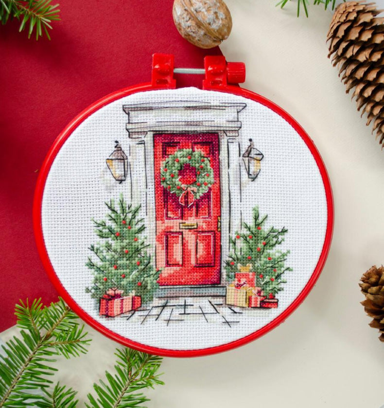 Cross-stitched design of a red door with a wreath, surrounded by Christmas decorations, on a white background.