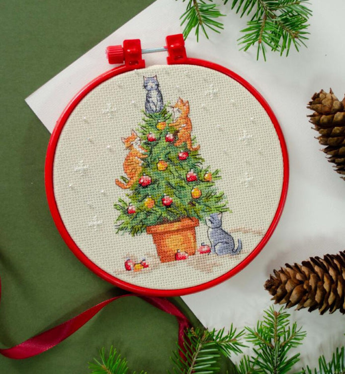 Embroidery hoop with a Christmas-themed cross-stitch design featuring cats around a decorated tree, surrounded by pine branches and cones.