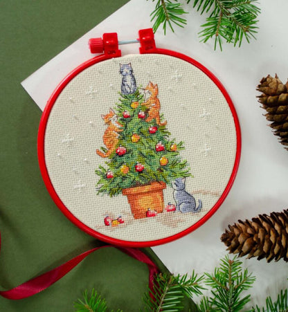 Embroidery hoop with a Christmas-themed cross-stitch design featuring cats around a decorated tree, surrounded by pine branches and cones.