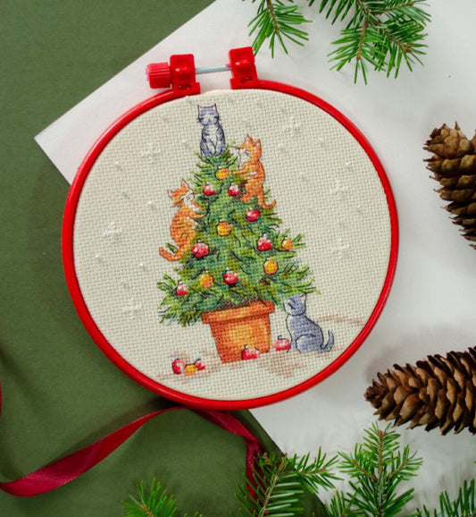 Embroidery hoop with a Christmas-themed cross-stitch design featuring cats around a decorated tree, surrounded by pine branches and cones.