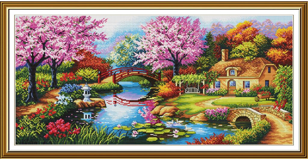 A framed cross-stitch or needlework-style landscape artwork showing a picturesque cottage beside a pond with pink flowering trees, a small bridge, lily pads, and colorful garden foliage. The piece is enclosed in a decorative gold-toned frame with a white mat.