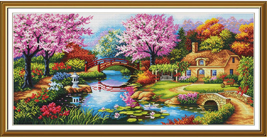 A framed cross-stitch or needlework-style landscape artwork showing a picturesque cottage beside a pond with pink flowering trees, a small bridge, lily pads, and colorful garden foliage. The piece is enclosed in a decorative gold-toned frame with a white mat.