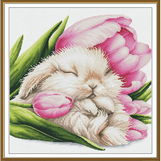 A framed needlepoint or cross-stitch style artwork showing a sleeping white bunny nestled among pink tulips and green leaves on a white canvas background, bordered by a gold frame.