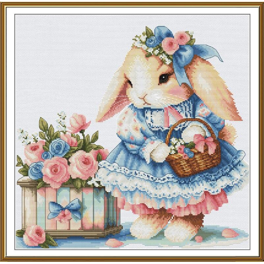 A framed cross-stitch style illustration of a bunny in a blue dress holding a basket of flowers, beside a matching flower box, with a white background and gold border.