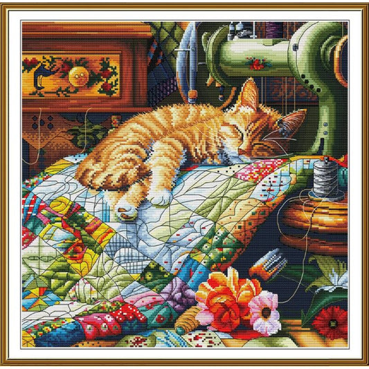 A framed needlework-style artwork showing a sleeping orange tabby cat resting on a colorful patchwork quilt, with a sewing machine, flowers, and other vintage domestic details in the background.