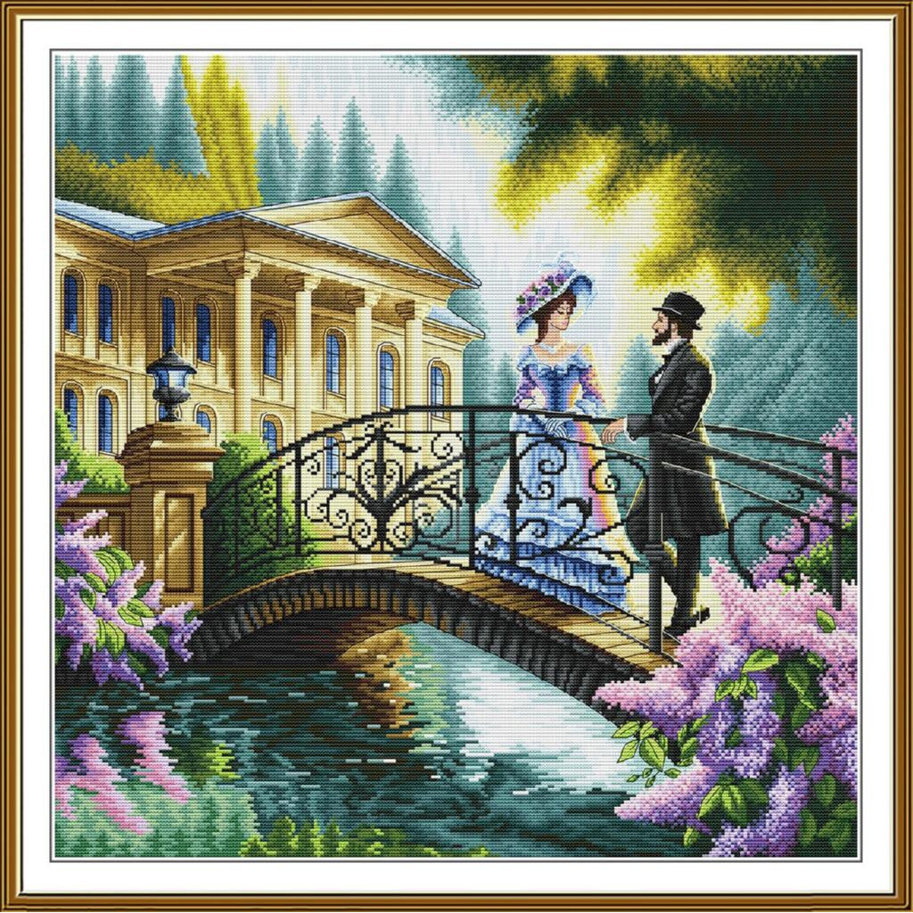 A framed cross-stitch or needlework-style art print depicting a romantic scene: a woman in a blue dress and hat stands beside a man in black on an ornate bridge over water, with a large classical building, trees, and purple flowers in the background.