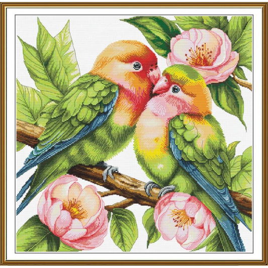 A framed cross-stitch style artwork showing two green lovebirds perched on a branch with pink flowers and green leaves on a white background, surrounded by a gold-toned frame.
