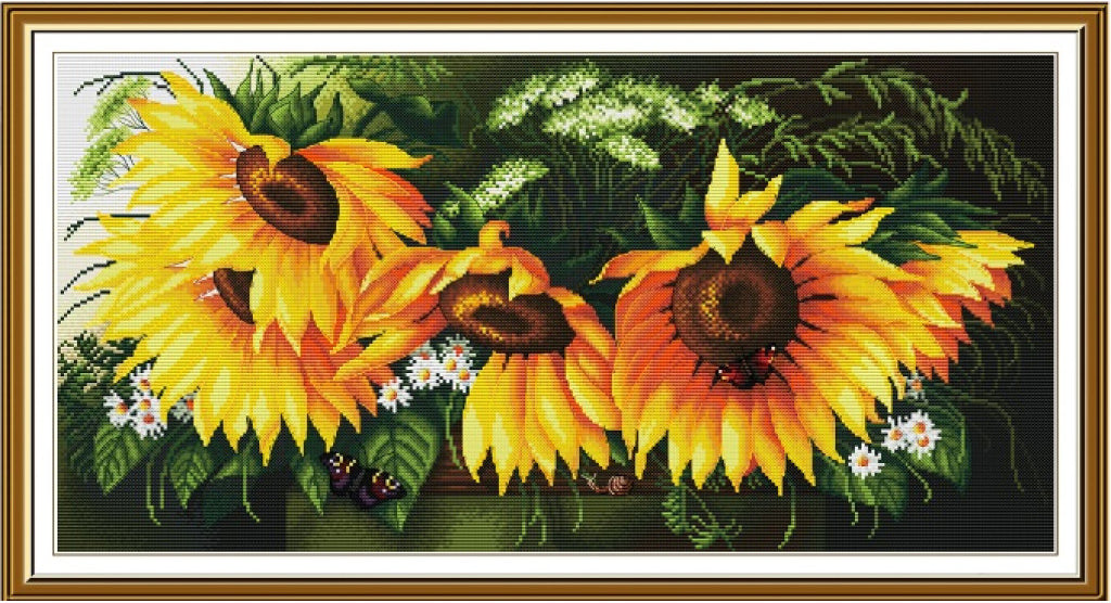 Framed cross-stitch or needlework-style floral artwork showing three large yellow sunflowers with dark brown centers, green leaves, and small white flowers, enclosed in a white mat and brown frame.