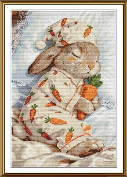 A framed needlepoint or cross-stitch style artwork showing a sleeping rabbit wearing a cream pajama set with an orange carrot print and a matching hat, holding an orange carrot, with a gold-toned frame and white mat border.
