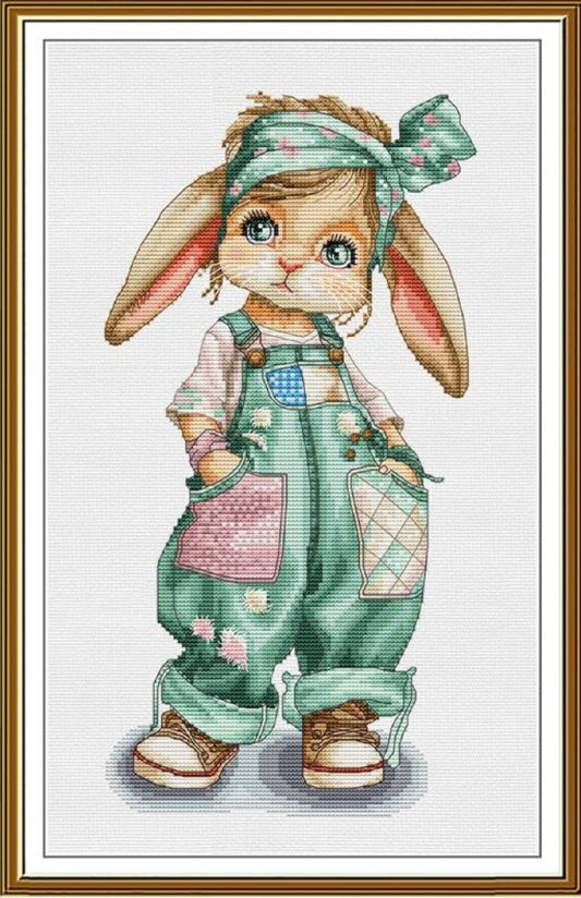 A framed cross-stitch style illustration of a cute bunny character wearing green overall-style clothing, a matching head bow, and brown shoes, shown against a white background with a gold-toned frame.