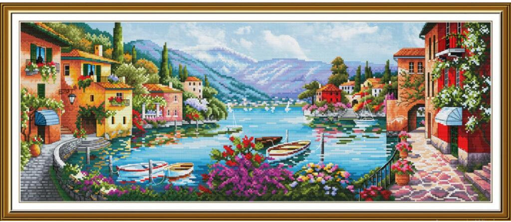 A framed cross-stitch or needlework-style scenic artwork depicting a lakeside village with colorful houses, boats, mountains, and flowers. The scene is enclosed in a decorative gold-toned frame and shown against a plain white background.