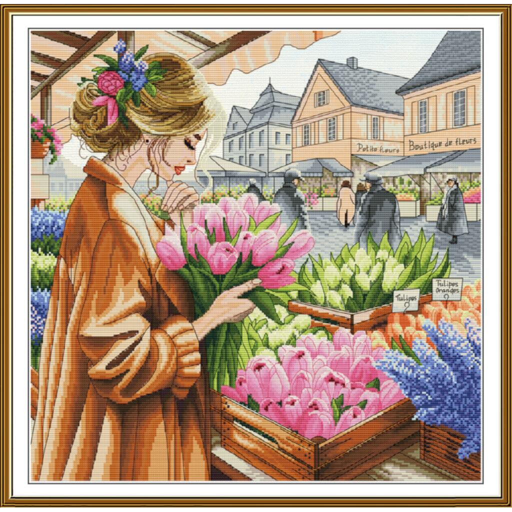 A framed cross-stitch or needlework-style art print depicting a woman in a brown coat holding pink tulips at a flower market, with buildings and other flowers in the background. The artwork is enclosed in a gold-toned frame.
