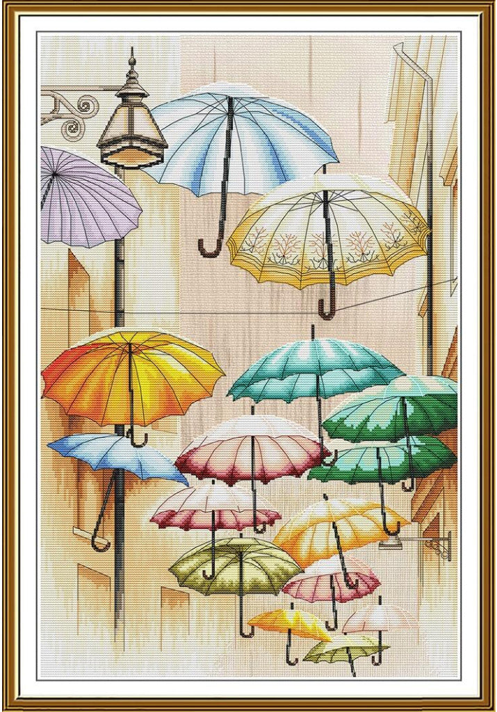 A framed needlepoint canvas featuring a whimsical street scene with multiple colorful umbrellas hanging in front of a beige building and a black lamppost. The canvas is framed in a gold-toned border with a white mat.